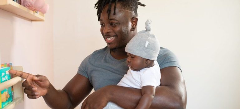 A man holding his baby and showing it something on a shelf.