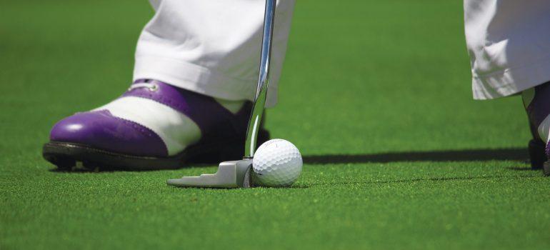 A shoe of the man, white golf ball and a golf club on the green grass of a gold field.