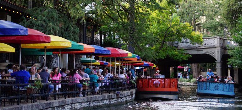 Riverwalk with restaurants full of people, a lot of greenery and two boats with tourists floating on the river.