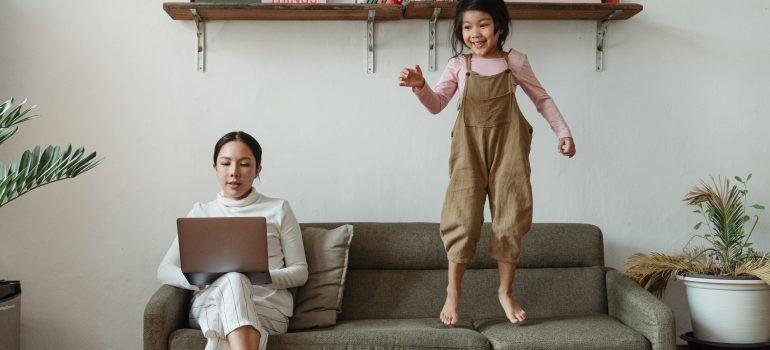 Mom holding a laptop sitting on a couch while little girls jumps on the couch