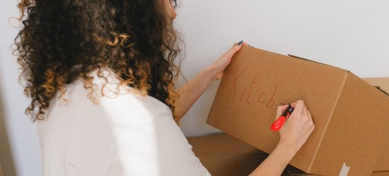 prepare for checking your inventory after an interstate move like this woman writing the word kitchen on the box 
