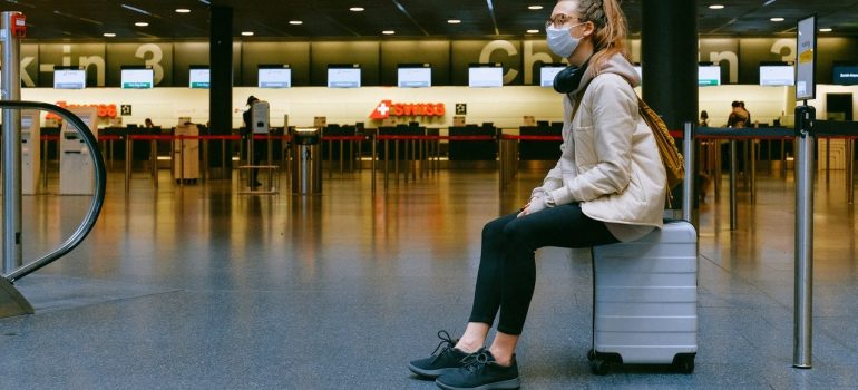a girl sitting on her suitcase in an airport while wearing a face mask