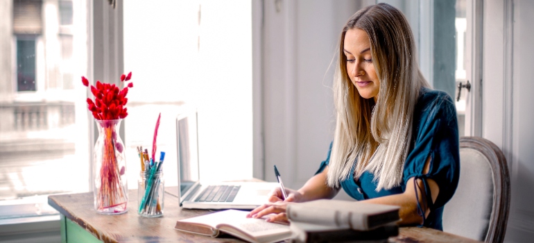 A woman writing something in a notebook while sitting at the desk that has a laptop and flowers on it.