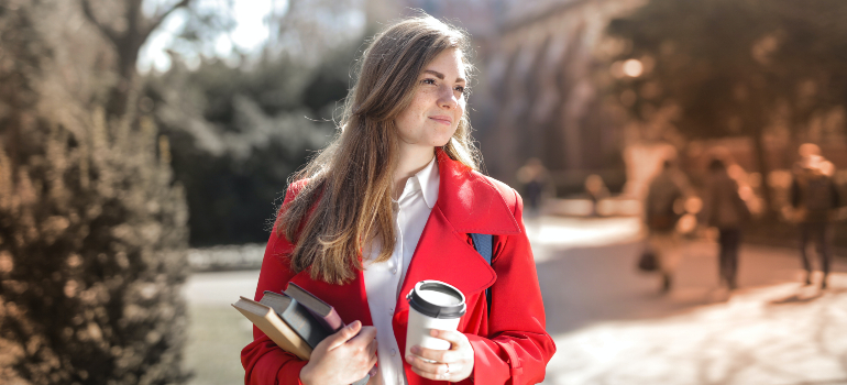A girl in a red coat holding a cup of coffee and books thinking about moving from South Houston to San Marcos